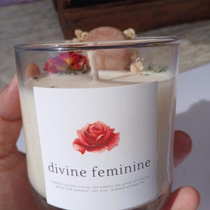 Op de afbeelding: Een heldere glazen kaars met een wit etiket met een rode roos en de woorden "divine feminine". De kaars is gevuld met witte was en versierd met gedroogde bloemen en een kleine steen. Het etiket bevat ook tekst.