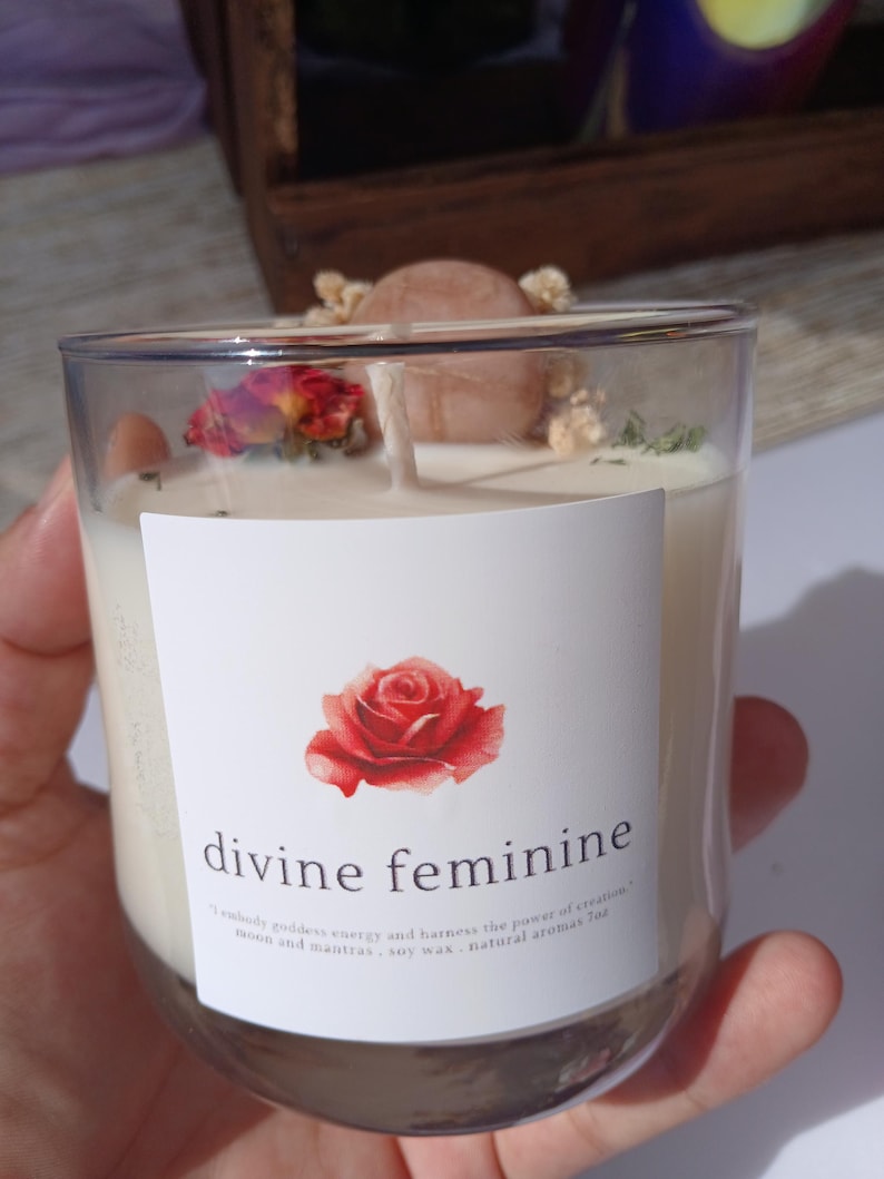 Op de afbeelding: Een heldere glazen kaars met een wit etiket met een rode roos en de woorden "divine feminine". De kaars is gevuld met witte was en versierd met gedroogde bloemen en een kleine steen. Het etiket bevat ook tekst.