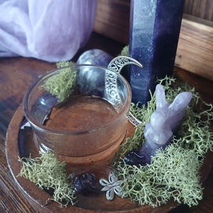 K&ouml;nnte beinhalten: Ein Holztablett mit einem durchsichtigen Glas-Votivkerzenhalter, einem geschnitzten Amethyst-Hasen und einem silbernen Halbmond-Anh&auml;nger. Das Tablett ist mit gr&uuml;nem Moos und kleinen Amethyststeinen verziert. Im Hintergrund steht ein hoher, dunkelvioletter Kristall.