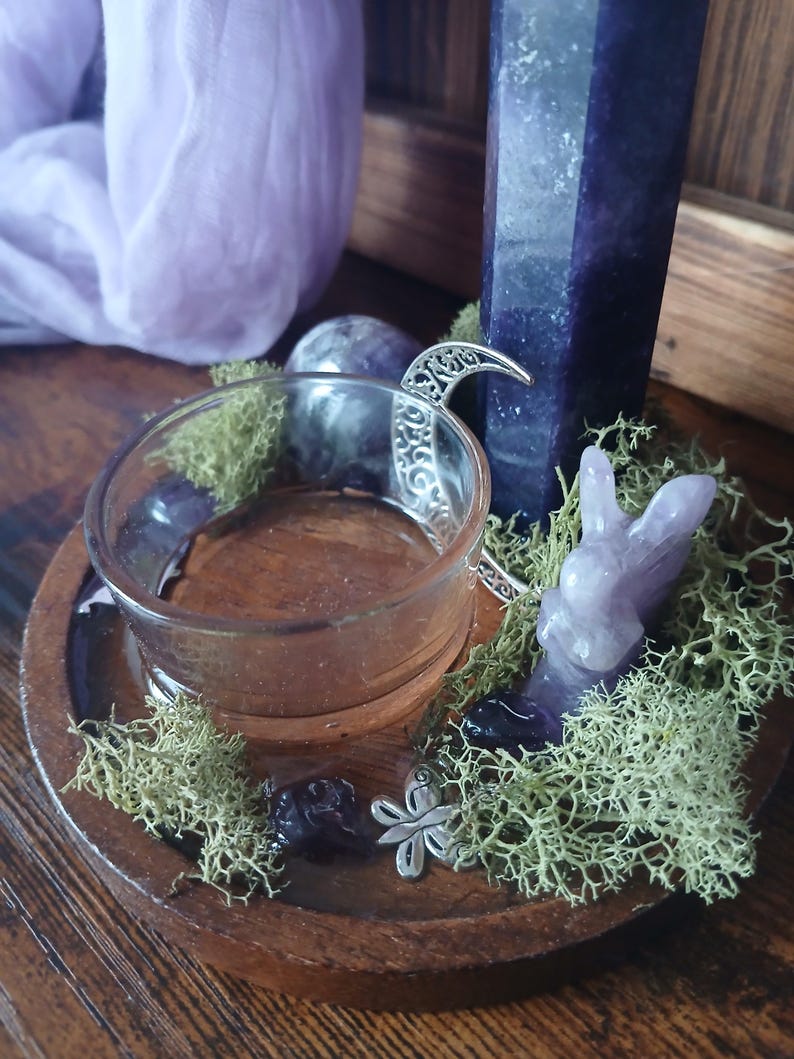 K&ouml;nnte beinhalten: Ein Holztablett mit einem durchsichtigen Glas-Votivkerzenhalter, einem geschnitzten Amethyst-Hasen und einem silbernen Halbmond-Anh&auml;nger. Das Tablett ist mit gr&uuml;nem Moos und kleinen Amethyststeinen verziert. Im Hintergrund steht ein hoher, dunkelvioletter Kristall.
