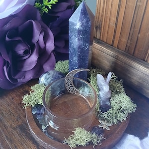 K&ouml;nnte beinhalten: Ein Holztablett mit einem klaren Glas, einem hohen lila Kristall und einer kleinen geschnitzten Kristallfigur. Das Tablett ist mit gr&uuml;nem Moos, kleinen Kristallen und einem dekorativen Halbmond verziert. Violette Blumen sind im Hintergrund.