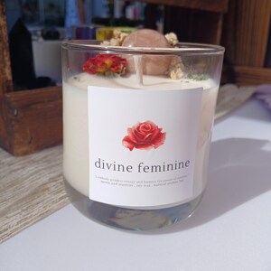 Op de afbeelding: Een heldere glazen kaars met witte was. De kaars is versierd met een rode roos, een roze steen en gedroogde bloemen. Het etiket leest "divine feminine" met een rozenillustratie.