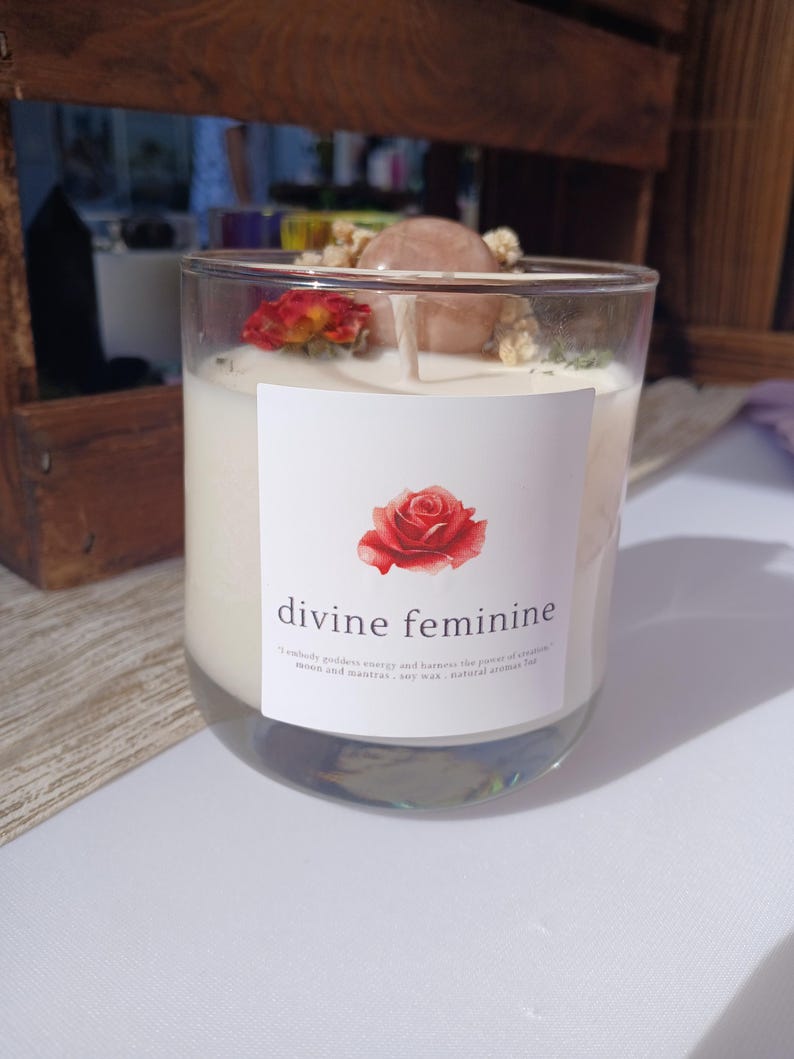 Op de afbeelding: Een heldere glazen kaars met witte was. De kaars is versierd met een rode roos, een roze steen en gedroogde bloemen. Het etiket leest "divine feminine" met een rozenillustratie.