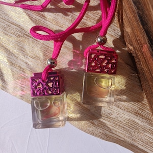 May include: Two small, clear, square glass bottles with a pink, decorative, openwork top and a pink cord. The bottles contain a light-coloured liquid. The cords are connected to a silver-coloured bead.