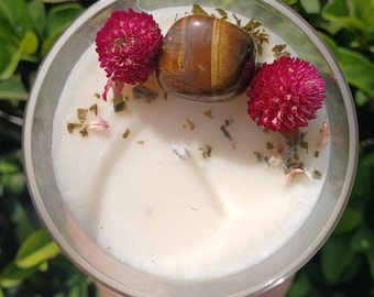 May include: A white wax candle in a clear glass container, topped with a polished brown and gold tiger's eye gemstone, two vibrant magenta globe amaranth flowers, and scattered dried green herbs surrounding a single centered cotton wick.