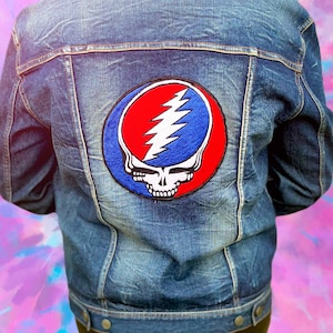 May include: A blue denim jacket with a large patch on the back featuring a red and blue Steal Your Face skull and lightning bolt design. The jacket has a classic button-up design and pockets.