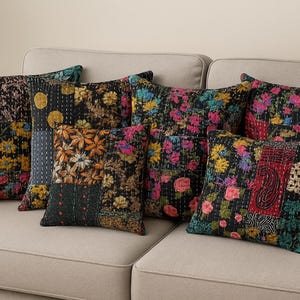 Puede incluir: Un conjunto de cojines decorativos con diseños florales de patchwork. Los cojines presentan una variedad de colores, incluyendo negro, rosa, amarillo y azul. Están colocados sobre un sofá beige, creando un ambiente acogedor.