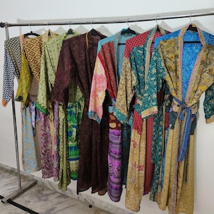 Op de afbeelding: Een verzameling kleurrijke, bedrukte badjassen die aan een metalen rek hangen. De badjassen hebben verschillende ontwerpen, waaronder geometrische vormen en bloempatronen, in tinten blauw, groen, rood en goud. Elke badjas heeft een bijpassende riem.