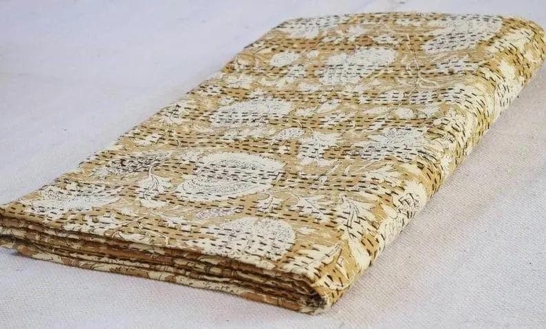 Może przedstawiać: Złożona, beżowo-biała kołdra w kwiaty. Kołdra ma powtarzający się kwiatowy wz&oacute;r z czarnymi przeszyciami. Kołdra jest złożona i ułożona w stos, pokazując krawędzie warstw. Tło to gładka, biała powierzchnia.