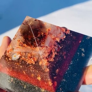 May include: A clear pyramid with layers of black, red, and white crystals and gold flakes. The pyramid is made of resin and is approximately 3 inches tall.