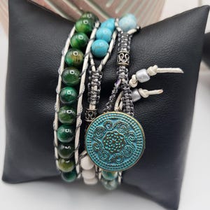 May include: A multi-strand bracelet featuring green, turquoise, and white beads. The bracelet includes a large, round, turquoise-colored button with a floral design. The bracelet is displayed on a black pillow.