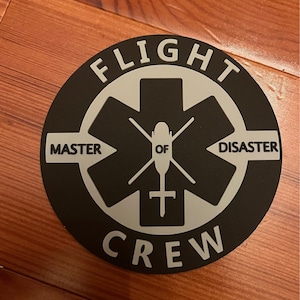 May include: Black and white round sticker with the text "FLIGHT CREW" in a circle around a white star of life with a helicopter inside. The text "MASTER OF DISASTER" is written on the points of the star.