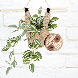 Könnte beinhalten: Ein gehäkelter Faultier-Pflanztopf, der an einer Holzleiste hängt, mit einer grün-weiß panaschierten Pflanze, die darin wächst. Das Faultier ist braun und hat schwarze Augen und einen süßen Ausdruck.