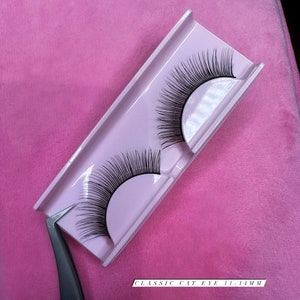 Könnte beinhalten: Zwei Paar schwarze falsche Wimpern in einem weißen Plastikbehälter mit dem Text "CLASSIC CAT EYE 11-14MM".