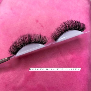 May include: Two pairs of black false eyelashes on a white plastic backing. The text "VOLUME DOLL EYE 11-13MM" is printed on a white label.