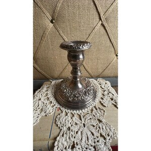 May include: A silver-toned metal candlestick holder featuring a wide, circular base decorated with embossed floral patterns. The stem is slender and cylindrical, leading to a flared top rim with a matching embossed leaf and floral design.