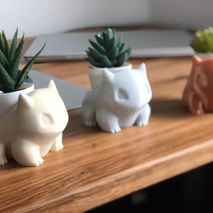 Könnte beinhalten: Drei kleine, dekorative Pflanzgefäße in Form von Zeichentrickfiguren, die jeweils eine kleine Sukkulentenpflanze enthalten. Die Pflanzgefäße sind in Creme, Grau und Orange gehalten und stehen auf einer Holzoberfläche. Die Pflanzen haben grüne Blätter.