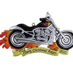 May include: A black and silver motorcycle ornament with flames coming from the back tire. The ornament has a yellow banner with the text "Merry Christmas Adam 2022".