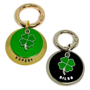 May include: Two gold and silver dog tags with a four-leaf clover charm. The gold tag has the name "RUPERT" engraved on it and the silver tag has the name "BILBO" engraved on it.