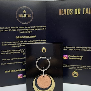 May include: A gold and copper pet ID tag with the text "I'm chipped" engraved on it. The tag is attached to a silver key ring. The tag is on a black card with the text "Heads or Tails Pet ID Tags" and social media handles.