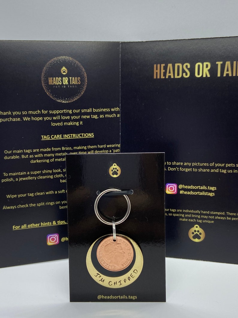 May include: A gold and copper pet ID tag with the text "I'm chipped" engraved on it. The tag is attached to a silver key ring. The tag is on a black card with the text "Heads or Tails Pet ID Tags" and social media handles.