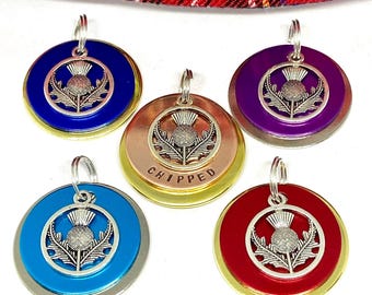 May include: Five round metal dog tags with a silver thistle design and a gold rim. The tags are in different colors: blue, purple, copper, turquoise, and red. One tag has the word "CHIPPED" engraved on it.