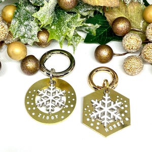 May include: Two gold-colored dog tags with snowflake charms. The tag on the left says "Ziggy" and the tag on the right says "Honey".
