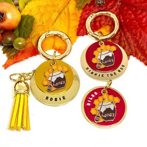 May include: Gold-colored keychains with round pendants. The pendants feature a honey pot design with the word "honey" and the names "Rosie", "Winnie the Woo", and "Bilbo". One keychain has a yellow tassel.