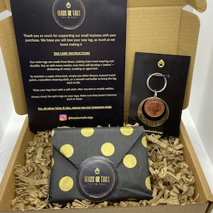 May include: A black and gold gift box with a black and gold polka dot envelope inside. The box contains a small brass pet ID tag with a dog paw print on it. The tag is attached to a silver key ring. The box is filled with brown packing peanuts. The box has a label that says "Heads or Tails Pet ID Tags".