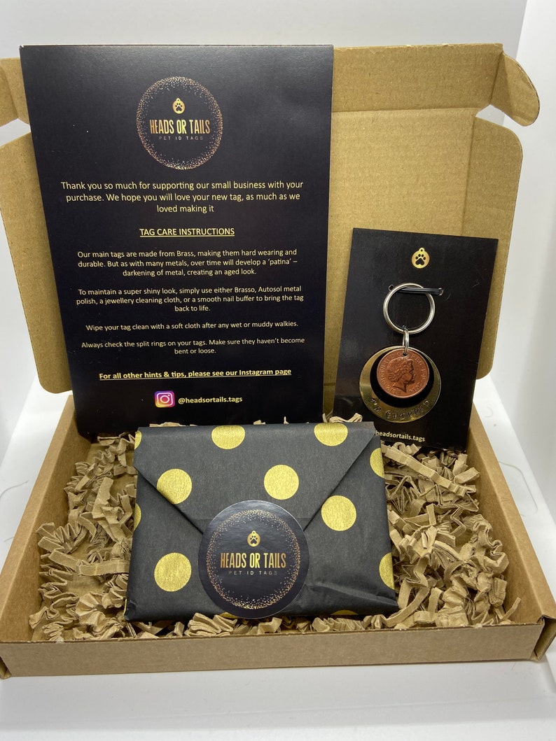 May include: A black and gold gift box with a black and gold polka dot envelope inside. The box contains a small brass pet ID tag with a dog paw print on it. The tag is attached to a silver key ring. The box is filled with brown packing peanuts. The box has a label that says "Heads or Tails Pet ID Tags".