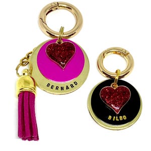 May include: Two gold-tone dog tag keychains with a red glitter heart charm. The keychain on the left has a pink background with the name "BERNARD" written in black. The keychain on the right has a black background with the name "BILBO" written in black. The keychain on the left has a pink tassel.