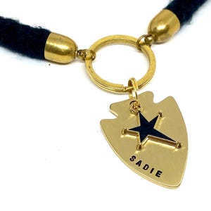 May include: Gold-tone keychain with a black tassel, a round key ring, and a gold arrow-shaped charm with a black star and the name "SADIE" engraved on it.