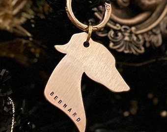 May include: A gold-colored metal keychain with a silhouette of a dog's head. The word "BERNARD" is engraved on the dog's head. The keychain is attached to a gold-colored ring.