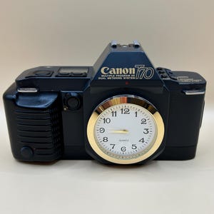 May include: A black Canon T70 camera-shaped clock with a gold-colored clock face. The clock face has black numbers and hands. The text "Canon T70" and "MULTIPLE PROGRAM AE DUAL METERING SYSTEM" are visible on the camera body.