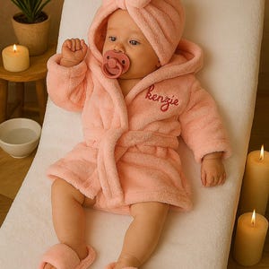 Peut inclure: Un b&eacute;b&eacute; v&ecirc;tu d'un peignoir &agrave; capuche rose clair, de chaussons assortis et d'un bonnet turban, avec une sucette dans la bouche. Le peignoir est brod&eacute; du nom "Kenzie". Le texte "Custom Baby Shower Gift Set" est en haut.