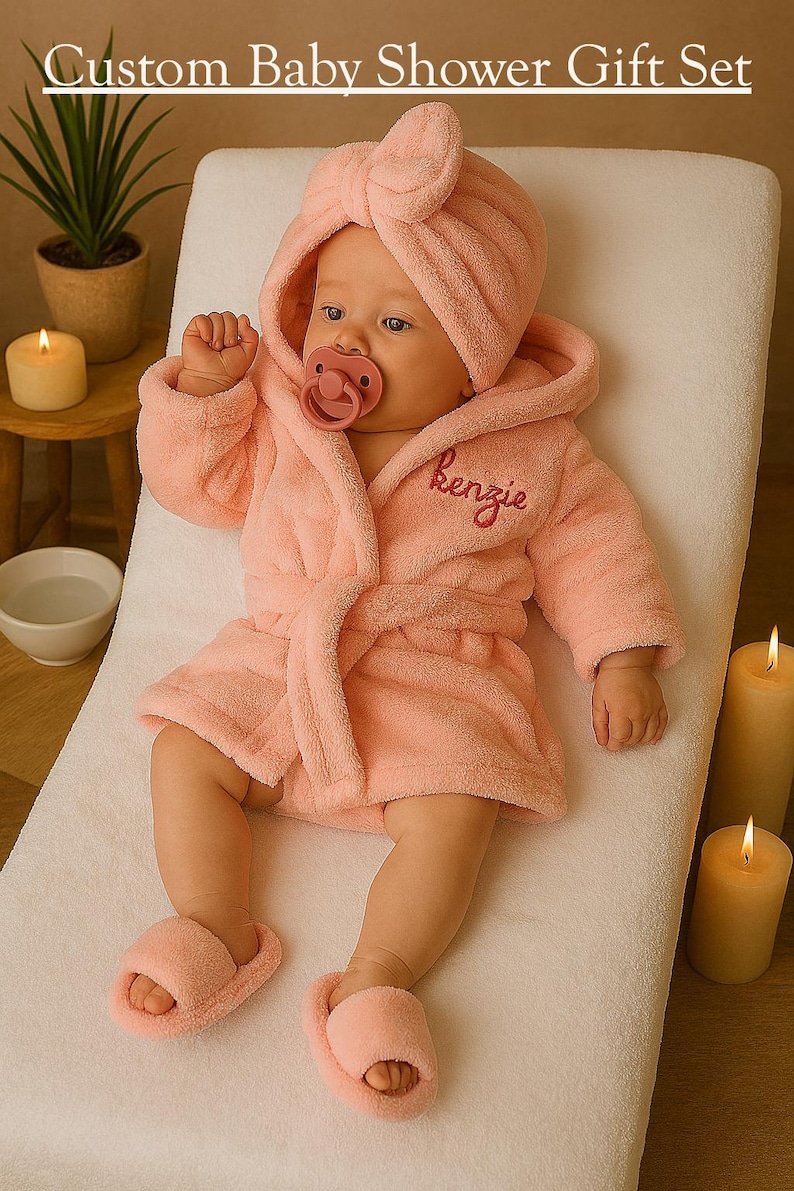 Peut inclure: Un b&eacute;b&eacute; v&ecirc;tu d'un peignoir &agrave; capuche rose clair, de chaussons assortis et d'un bonnet turban, avec une sucette dans la bouche. Le peignoir est brod&eacute; du nom "Kenzie". Le texte "Custom Baby Shower Gift Set" est en haut.