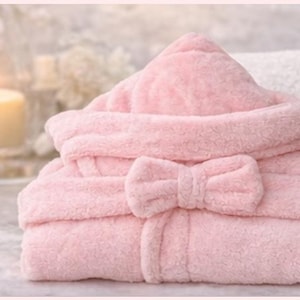 Peut inclure: Un peignoir &agrave; capuche rose clair avec une fermeture &agrave; n&oelig;ud papillon assortie. Le peignoir en peluche est pli&eacute; avec soin, mettant en valeur sa texture et son design. L'image &eacute;voque le confort et la d&eacute;tente, pour le spa ou le bain.