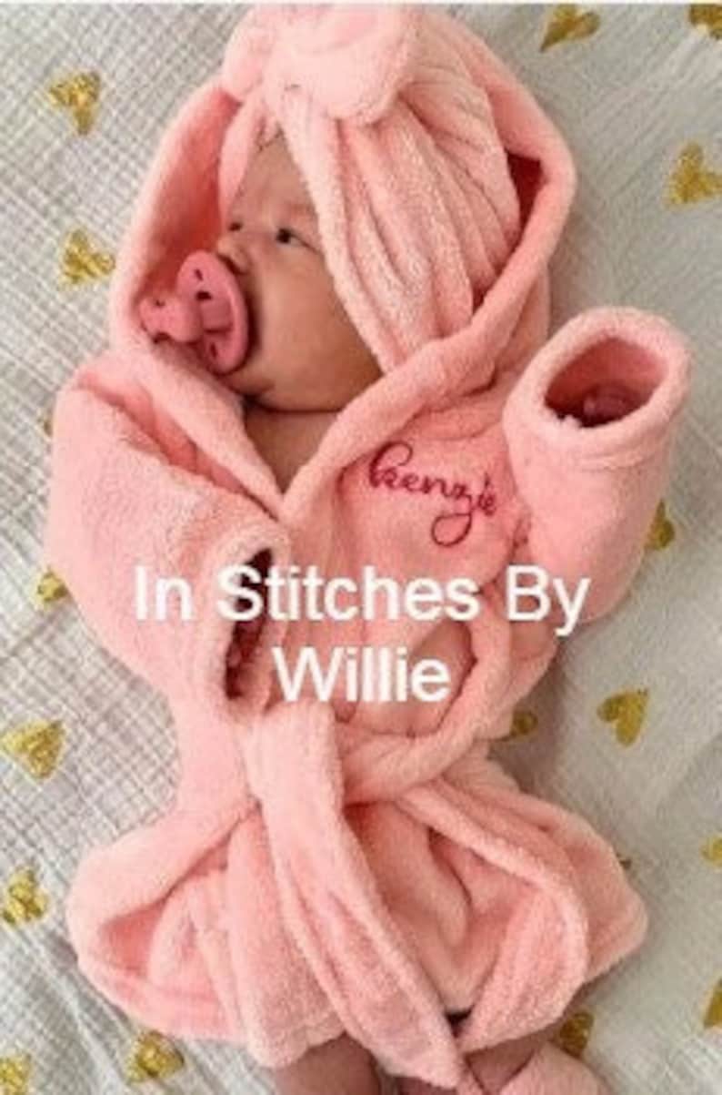 Peut inclure: Un b&eacute;b&eacute; portant un peignoir &agrave; capuche rose clair et une sucette. Le peignoir est brod&eacute; du nom "Renz's". Le texte "In Stitches By Willie" est &eacute;galement visible.