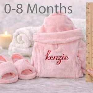 Peut inclure: Ensemble de peignoir et chaussons roses pour b&eacute;b&eacute;. Le peignoir est brod&eacute; du nom "kenzie" en &eacute;criture rouge. L'image comprend &eacute;galement une r&egrave;gle, indiquant la taille des articles. Le texte "0-8 Months" est en haut.