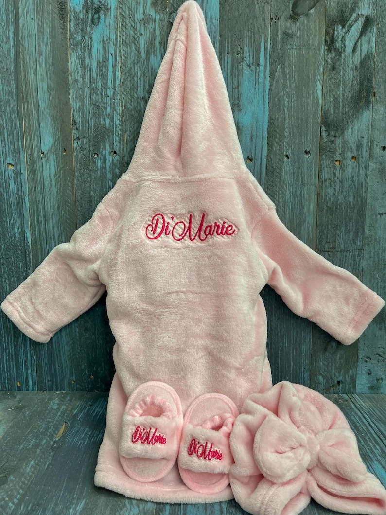 Peut inclure: Ensemble de robe en peluche rose clair avec pantoufles et bonnet assortis. La robe et les pantoufles sont brod&eacute;es du nom "Di Marie" en &eacute;criture cursive. L'ensemble est pr&eacute;sent&eacute; sur une surface en bois bleu.