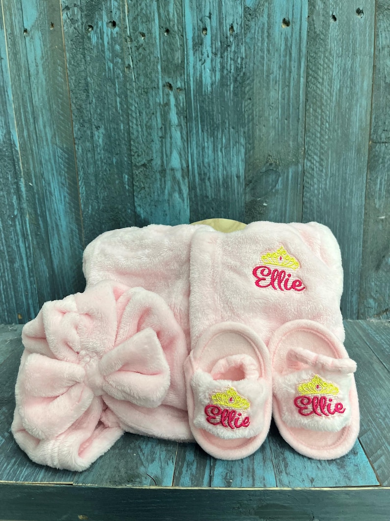 Peut inclure: Ensemble de spa rose en peluche comprenant un bandeau avec un n&oelig;ud, un peignoir et une paire de chaussons. Le peignoir et les chaussons sont brod&eacute;s d'une couronne et du nom "Ellie".