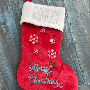 May include: Red Christmas stocking with white faux fur cuff, embroidered with the name "Raquel" in silver glitter thread, snowflakes, and the phrase "Merry Christmas" in teal thread.