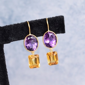 May include: A pair of gold earrings with purple and orange gemstones. The earrings feature a teardrop-shaped purple gemstone and a square-shaped orange gemstone.