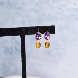 May include: A pair of gold earrings with a purple oval gemstone and a smaller, rectangular, yellow gemstone hanging below.
