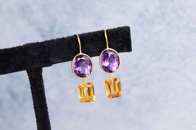 May include: A pair of gold earrings with purple and orange gemstones. The earrings feature a teardrop-shaped purple gemstone and a square-shaped orange gemstone.