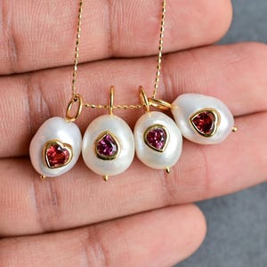May include: Four gold necklaces with white pearl pendants, each featuring a heart-shaped gemstone. The gemstones are either red or pink. The necklaces are displayed on a hand, with a silver ring visible.