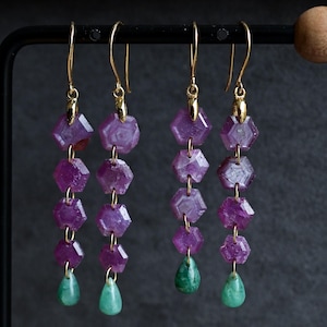 May include: Dangling earrings with gold hooks feature a series of faceted, hexagonal purple gemstones, connected by gold links. Each earring ends with a teardrop-shaped green gemstone. The earrings are displayed against a dark gray background.