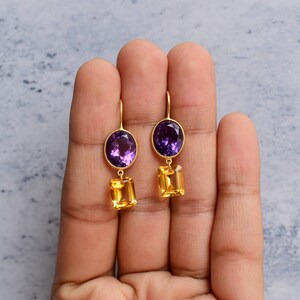 May include: A pair of gold earrings with purple and yellow gemstones. The earrings feature a round purple gemstone at the top and a rectangular yellow gemstone dangling below.