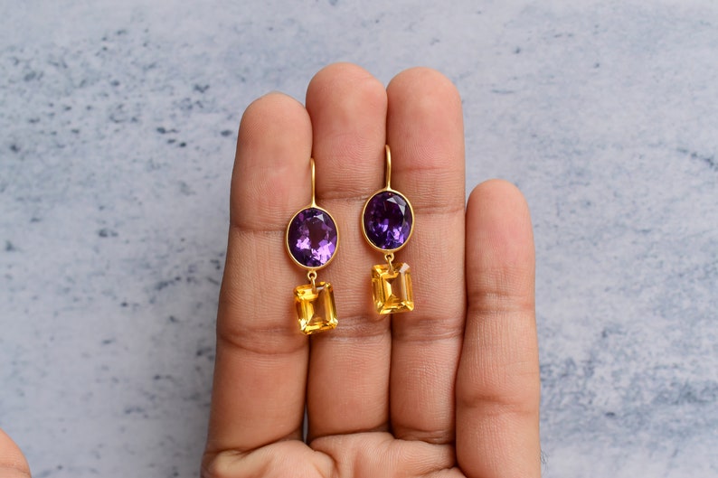 May include: A pair of gold earrings with purple and yellow gemstones. The earrings feature a round purple gemstone at the top and a rectangular yellow gemstone dangling below.