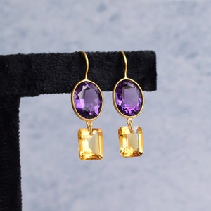May include: A pair of gold earrings with a purple oval-shaped gemstone and a smaller, square-shaped yellow gemstone dangling below.